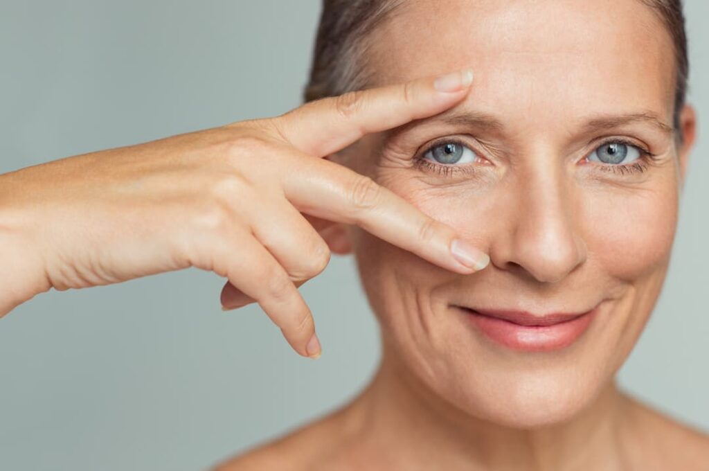 Botox treatments - Portrait of smiling senior woman with perfect skin showing victory sign near eye on grey background.