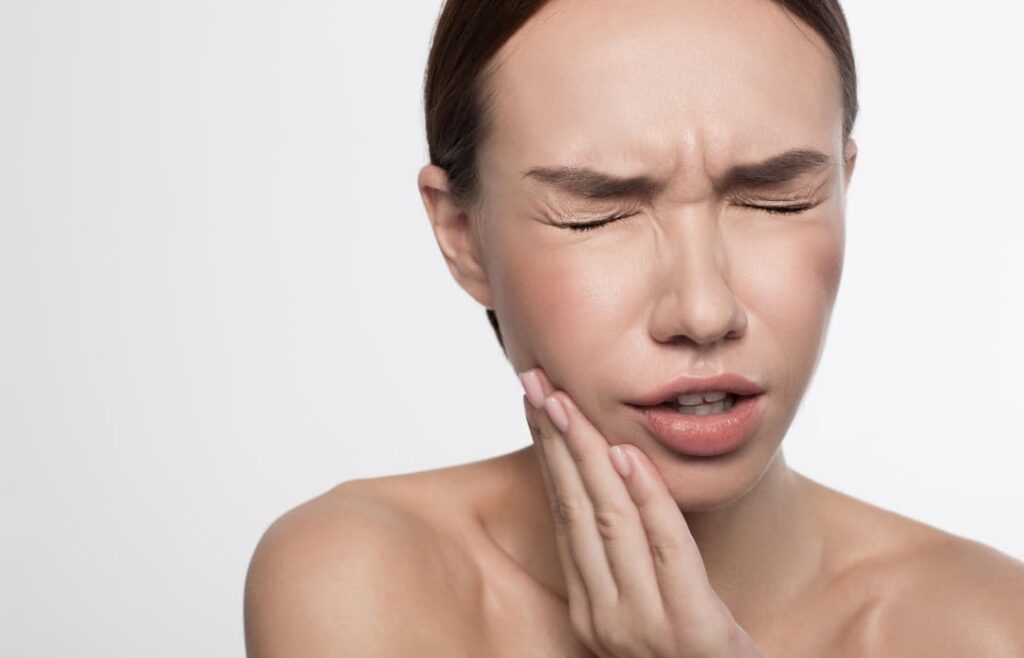 Botox for TMJ and Bruxism - Portrait of unhappy girl is standing and holding her hand on cheek with closed eyes while wincing her face in terrible toothache.