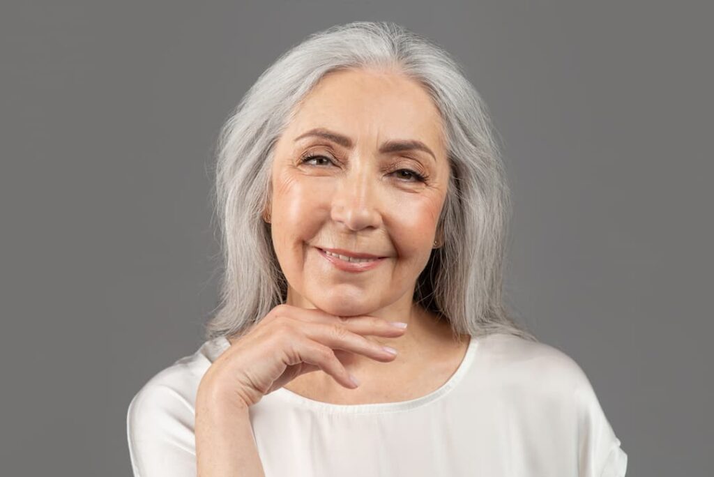 Radiesse & Hyperdilute Radiesse treatments - Beauty portrait of senior woman touching her face, looking at camera and smiling on grey studio background