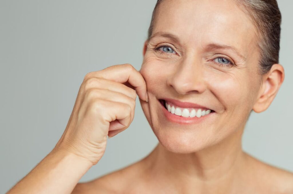 Sculptra teatments - Senior woman pulling cheeks to feel softness and looking at camera.