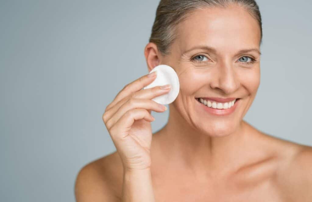 Skin Health - Healthy mature woman removing makeup from her face with cotton pad isolated on grey background. Beauty portrait of happy woman cleaning skin and looking at camera.